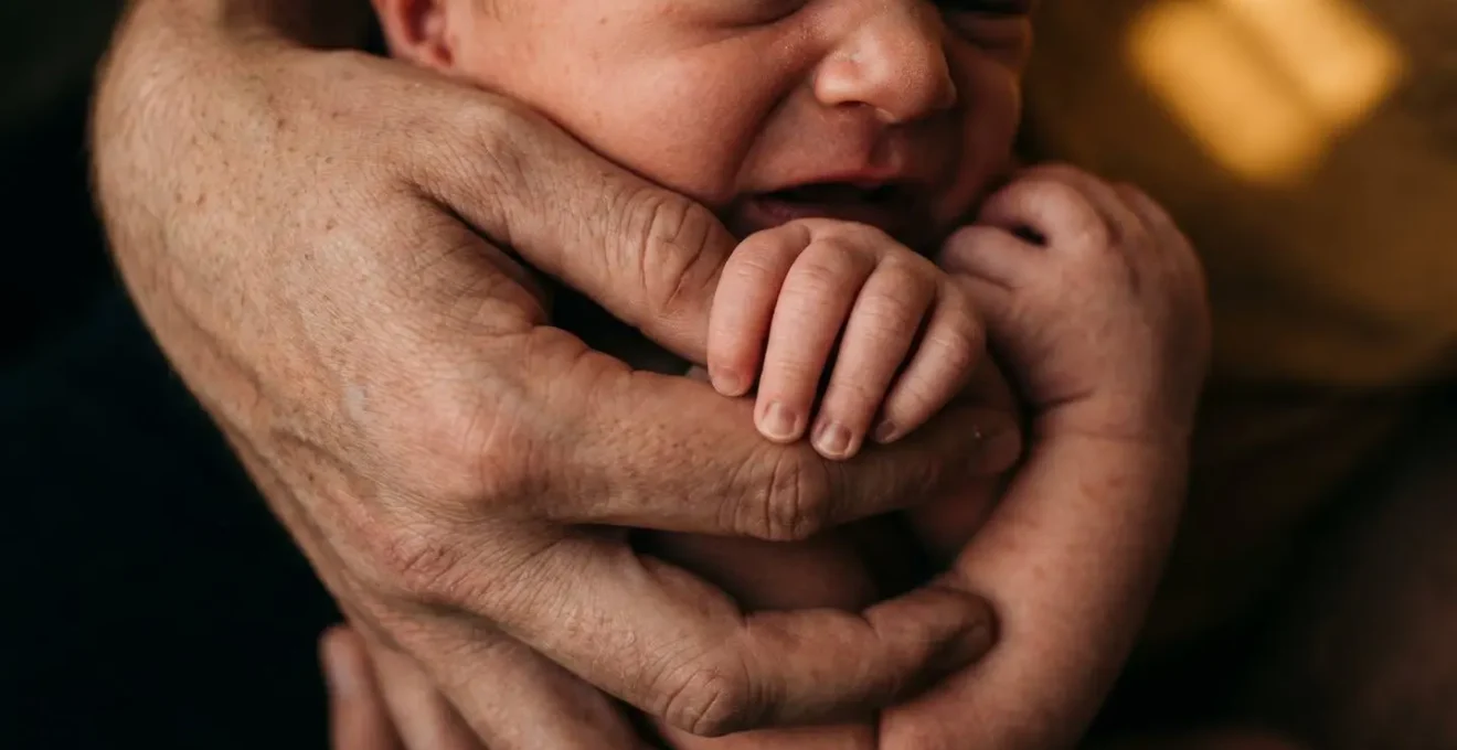 Gros plan sur les mains d'un parent tenant doucement un nourrisson en pleurs dans une lumière tamisée de fin de journée