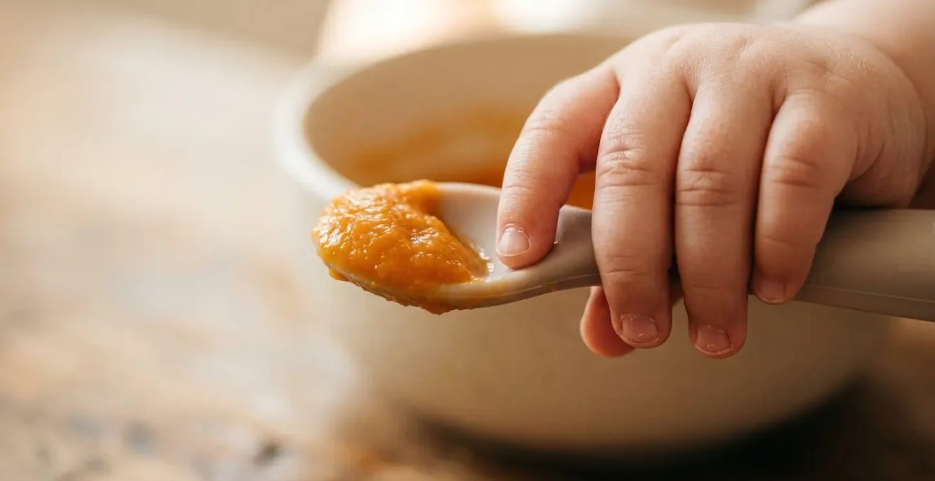 Gros plan sur la main potelée d'un bébé explorant une purée de légumes orange avec une cuillère adaptée